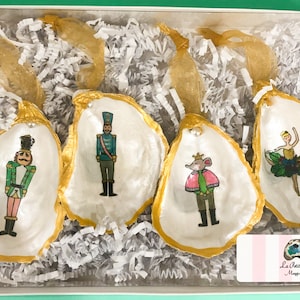 May include: Set of four oyster shell ornaments painted with characters from The Nutcracker. The ornaments feature a nutcracker soldier, a mouse king, a ballerina, and a second nutcracker soldier. The shells are painted white with gold trim and are packaged in a white box with a pink and white striped label that reads "La Petite Shell - Maggie Grimes".