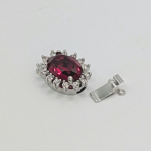 May include: A silver-toned jewelry clasp featuring a large, oval-shaped deep red gemstone at the center, surrounded by a halo of small, clear, sparkling stones. A separate, plain silver-toned metal hook component lies next to the main clasp.