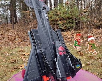 May include: A black toy fighter jet with red cockpit windows, silver missile details, and red engine exhausts. The aircraft features a Cobra logo on the wing and is displayed in a vertical position on a black stand, accompanied by two small gingerbread man stickers.
