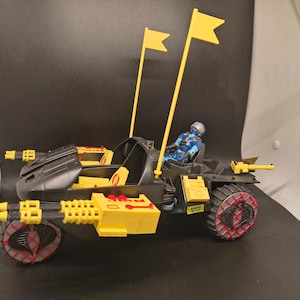 Op de afbeelding: Een zwart en geel speelgoedvoertuig met een blauwe figuur op de bestuurdersstoel. Het voertuig heeft twee gele vlaggen, rood-grijze wielen en gele machinegeweren. Het speelgoed staat tegen een zwarte achtergrond.