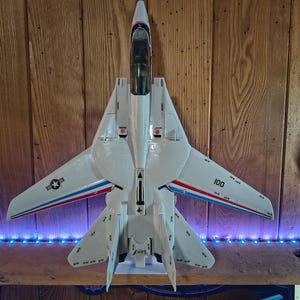 May include: A white model fighter jet with swept-back wings, featuring red and blue accent stripes along the wings, a black cockpit canopy, and the number 100 printed on the right wing. A circular military insignia is visible on the left wing.