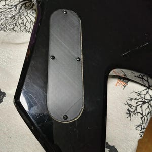 May include: Close-up of a black electric guitar body with a rectangular, textured gray plate secured by four screws. The guitar has a glossy finish and shows signs of wear.