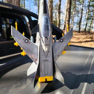 Peut inclure: Un avion jouet gris et blanc avec des accents jaunes, des ailes et des missiles. Le jouet a un cockpit transparent et présente un emblème d'étoile sur les ailes. L'avion est posé sur une surface sombre avec un arrière-plan flou d'arbres.