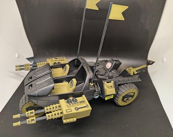 May include: A black and olive green toy vehicle with two yellow flags, two seats, and multiple weapon attachments. The toy has a military theme and is designed with intricate details, including a star emblem on the wheel.