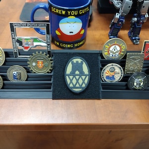 May include: A black display case with 10 slots for displaying military challenge coins. The case is filled with various coins, including a coin with the words "Operation Spartan Shield" and a coin with the words "STC".