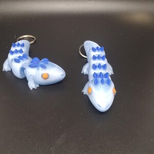 May include: Two blue and white lizard keychains with orange eyes and blue spikes. The lizards are posed in a walking position.