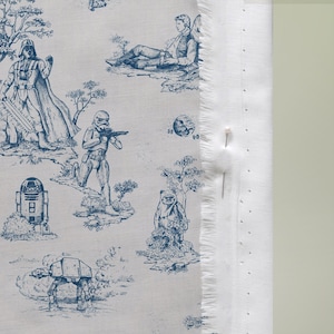 May include: White fabric with a blue toile pattern featuring Star Wars characters and scenes. The design includes Darth Vader, Stormtroopers, R2-D2, and an AT-AT walker. The fabric has a raw edge with a pin.