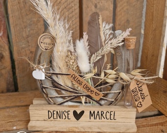 May include: A decorative horseshoe arrangement on a wooden base labeled Denise and Marcel. It features dried grasses, a brown feather, a glass vial, and a tag reading Alles Liebe. A small white heart and a circular tag reading Du & Ich are attached.