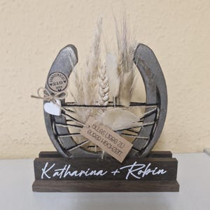 Könnte beinhalten: Rustikale Hochzeitsdekoration aus Holz mit Hufeisen-Design, gefüllt mit Trockenblumen und Federn. Auf der Basis stehen die Namen "Katharina + Robin". Ein Anhänger trägt die Aufschrift "Alles Liebe zu eurer Hochzeit".