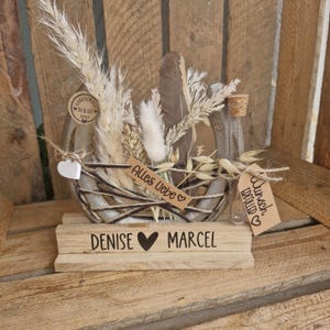 Könnte beinhalten: Rustikaler Holz-Mittelstück mit einem geflochtenen Korb, gefüllt mit getrockneten Blumen, Federn und kleinen Glasflaschen. Die Basis ist mit "DENISE" und "MARCEL" graviert, getrennt durch ein Herzsymbol. Tags lesen "Alles Liebe" und "Wunsch Erfüllt".