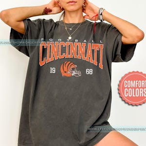 May include: A dark gray t-shirt with the text "FOOTBALL CINCINNATI 19 68" and a brown and orange football helmet graphic.