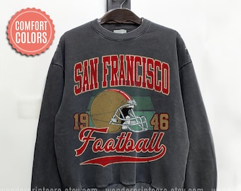 Può includere: Felpa grigio scuro con un design vintage di San Francisco Football. Il design presenta un casco da football, le parole "San Francisco", "1946" e "Football" in lettere rosse e dorate. La felpa è appesa a una gruccia nera.