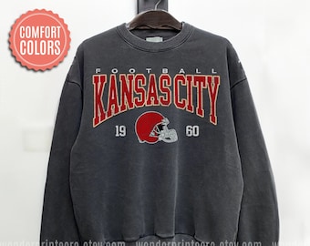 May include: A dark gray sweatshirt with a vintage-style graphic print of "FOOTBALL KANSAS CITY 19 60" in red and yellow lettering with a red football helmet graphic.