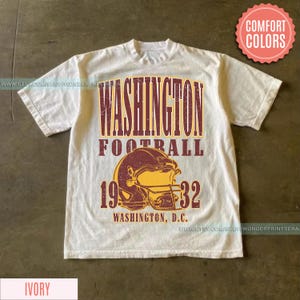 May include: Ivory-colored t-shirt featuring a distressed graphic. The design includes the text "WASHINGTON FOOTBALL" above a football helmet and the numbers "1932". The words "WASHINGTON, D.C." are printed below the helmet.