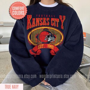 Pode incluir: Uma camisola azul marinho com um gr&aacute;fico de estilo vintage de um capacete de futebol e o texto "FOOTBALL KANSAS CITY EST. 1960".