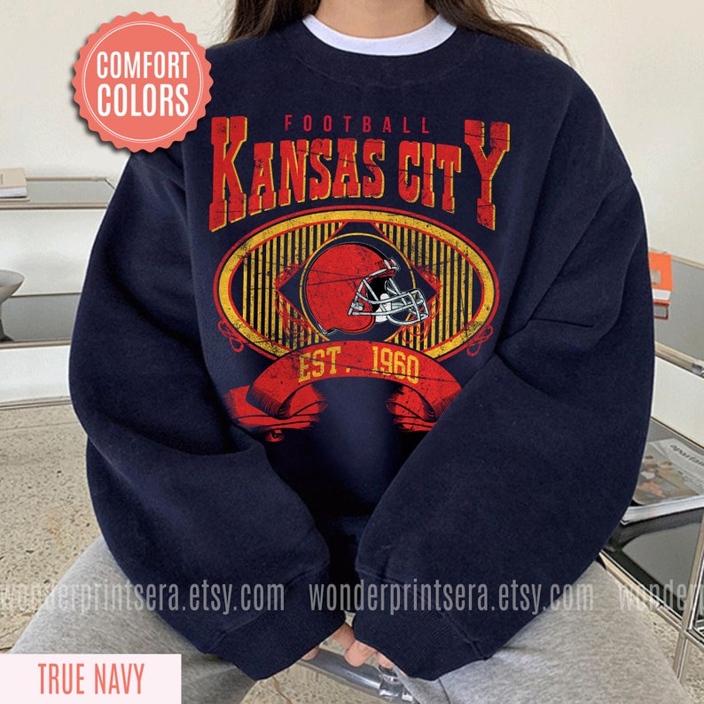 Pode incluir: Uma camisola azul marinho com um gr&aacute;fico de estilo vintage de um capacete de futebol e o texto "FOOTBALL KANSAS CITY EST. 1960".