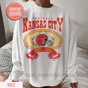 Pode incluir: Camisola branca com um gr&aacute;fico de estilo vintage de um capacete de futebol e o texto "FOOTBALL KANSAS CITY EST. 1960".