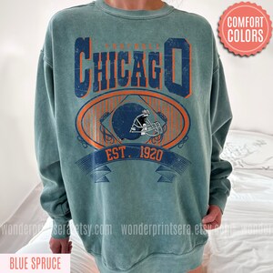 May include: A teal blue sweatshirt with a vintage-style graphic of a football helmet and the text "FOOTBALL CHICAGO EST. 1920".