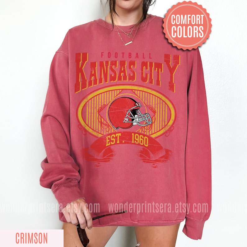 Pode incluir: Camisola de cor vermelha carmim com um gr&aacute;fico de estilo vintage de um capacete de futebol e o texto "FOOTBALL KANSAS CITY EST. 1960".