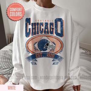 May include: White sweatshirt with a vintage-style graphic design featuring the text "FOOTBALL CHICAGO EST. 1920" and a football helmet.
