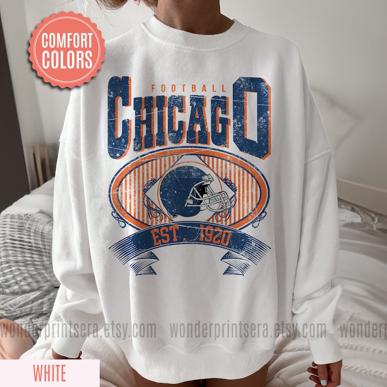May include: White sweatshirt with a vintage-style graphic design featuring the text "FOOTBALL CHICAGO EST. 1920" and a football helmet.