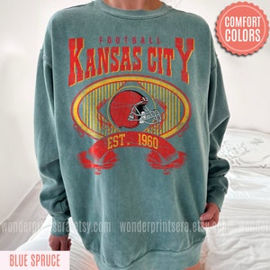 Pode incluir: Uma camisola azul-esverdeada com um gr&aacute;fico de estilo vintage de um capacete de futebol e o texto "FOOTBALL KANSAS CITY EST. 1960".