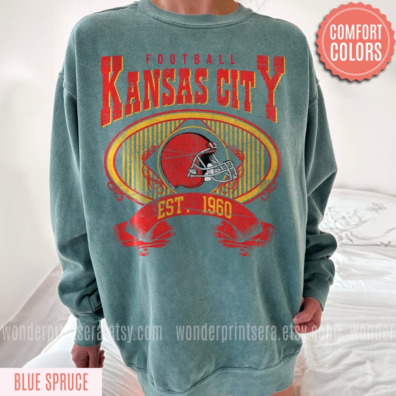 Pode incluir: Uma camisola azul-esverdeada com um gr&aacute;fico de estilo vintage de um capacete de futebol e o texto "FOOTBALL KANSAS CITY EST. 1960".