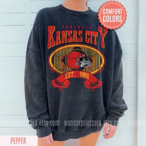 Pode incluir: Uma camisa de frio preta com um design gr&aacute;fico de estilo vintage que apresenta o texto "Football Kansas City Est. 1960" e um capacete de futebol vermelho e branco. O gr&aacute;fico &eacute; cercado por um oval com listras amarelas e pretas.
