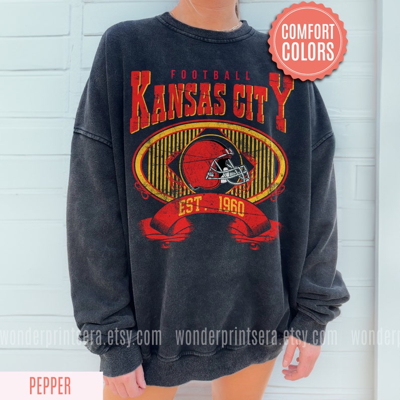Pode incluir: Uma camisa de frio preta com um design gr&aacute;fico de estilo vintage que apresenta o texto "Football Kansas City Est. 1960" e um capacete de futebol vermelho e branco. O gr&aacute;fico &eacute; cercado por um oval com listras amarelas e pretas.