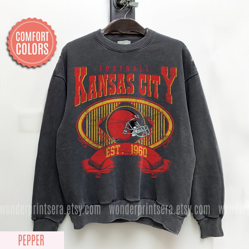 Pode incluir: Uma camisola cinzenta escura com um gr&aacute;fico de estilo vintage de um capacete de futebol e o texto "FOOTBALL KANSAS CITY EST. 1960".