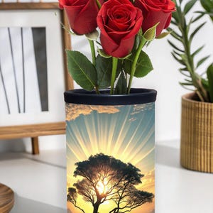 Könnte beinhalten: Eine zylindrische Vase mit schwarzem Rand, die einen Strauß leuchtend roter Rosen enthält. Die Vase zeigt ein szenisches Design eines Baumes vor einem Sonnenuntergang mit Sonnenstrahlen. Die Vase steht auf einer weißen Oberfläche.