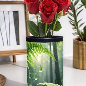 Può includere: Un vaso cilindrico con un design a scena di foresta, con felci e gocce d'acqua, che contiene un bouquet di rose rosse. Il vaso ha un bordo nero. Lo sfondo include un'opera d'arte incorniciata e una pianta in vaso.
