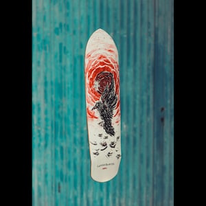 May include: A skateboard with a white surface featuring a red and black design. The design includes a swirling red pattern, a black raven, and the text "Lunda Boards". The skateboard has a rounded nose and tail.
