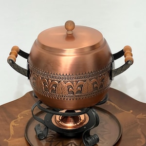 Può includere: Pentola per fonduta in rame con un pomello in legno sul coperchio e manici in legno. La pentola è su un supporto in metallo nero con un vassoio in rame.