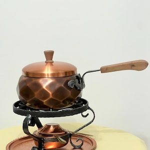 Könnte beinhalten: Ein Fonduetopf aus Kupfer mit einem Holzgriff und einem Deckel, der auf einem schwarzen Metallständer mit einem Kupfertablett steht. Der Topf hat eine gehämmerte Oberfläche und ein dekoratives Design.