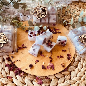 Puede incluir: Cuatro fundentes de cera transparentes con pétalos de rosa secos y una decoración de madera en forma de rosa. Los fundentes de cera están envasados en recipientes de plástico transparentes con un borde dorado. Los fundentes de cera están sobre una superficie de madera con pétalos de rosa secos esparcidos alrededor.