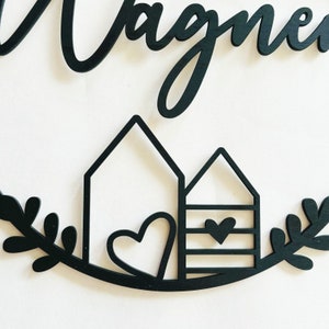 May include: Black wooden sign with the word "Wagner" in cursive script. The sign features two stylized houses with hearts and a leafy wreath.
