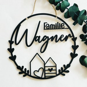 Op de afbeelding: Rond zwart houten bord met de tekst "Familie Wagner" en twee gestileerde huizen met harten. Het bord is versierd met een krans van bladeren.