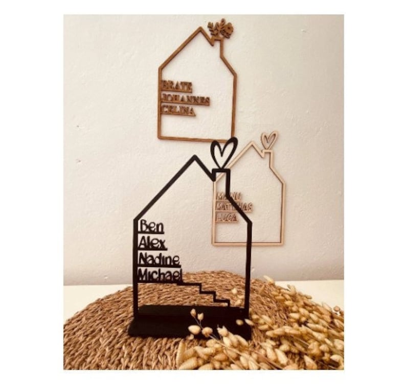 May include: Black wooden house-shaped sign with the names Ben, Alex, Nadine, and Michael written on it. The sign is standing on a woven straw surface.