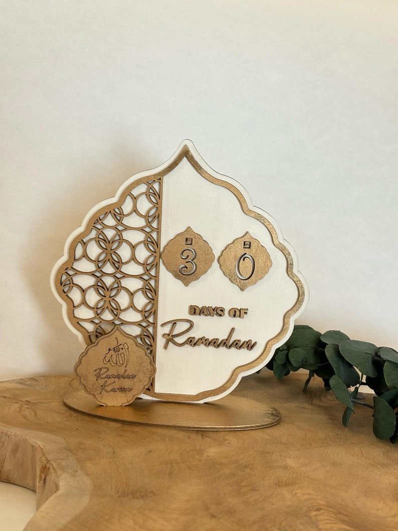 Pu&ograve; includere: Un calendario di conto alla rovescia in legno bianco con accenti dorati e tagli intricati. Il calendario presenta il testo "Days of Ramadan" e i numeri "30". &Egrave; incluso anche un piccolo cerchio dorato con il testo "Ramadan Kareem".