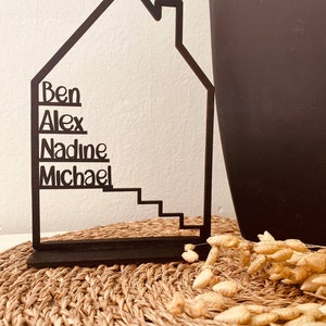 May include: Black metal house-shaped silhouette with the names Ben, Alex, Nadine, and Michael written on it. The house has a heart-shaped chimney and a set of stairs. The house is sitting on a woven mat.