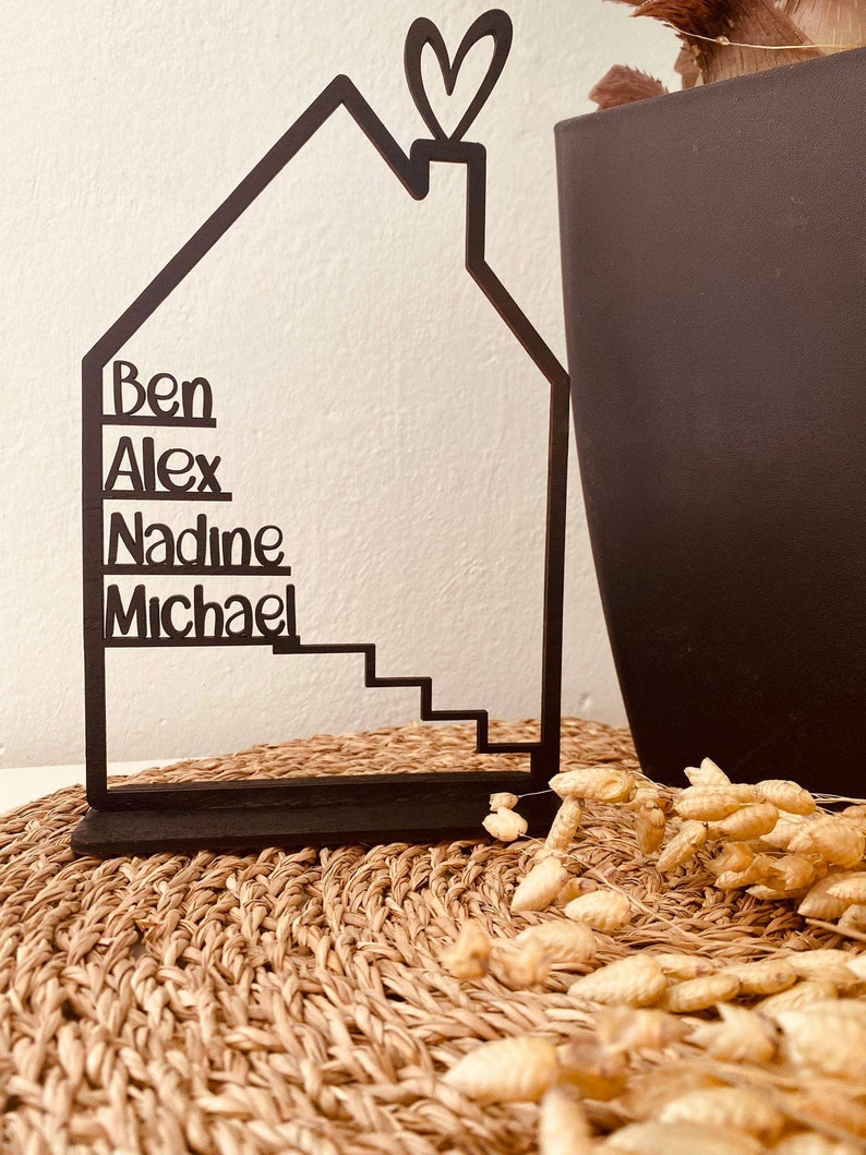 May include: Black metal house-shaped silhouette with the names Ben, Alex, Nadine, and Michael written on it. The house has a heart-shaped chimney and a set of stairs. The house is sitting on a woven mat.