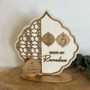 Pu&ograve; includere: Un calendario di conto alla rovescia in legno bianco e oro con un design decorativo. Il calendario presenta due cerchi dorati con i numeri 30 e 0, e il testo "DAYS OF RAMADAN". C'&egrave; un cerchio dorato pi&ugrave; piccolo con il testo "Ramadan Kareem" e un simbolo arabo.
