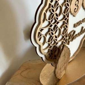 Pu&ograve; includere: Un porta numero da tavolo in legno con un design decorativo tagliato al laser. Il numero "3" &egrave; visualizzato sul davanti, insieme al testo "Giorni fino al nostro matrimonio".