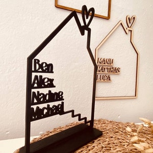 May include: Black wooden house-shaped sign with the names Ben, Alex, Nadine, and Michael written vertically on the front. The sign has a heart-shaped cutout at the top.