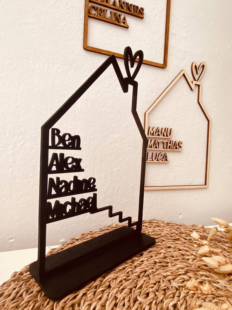 May include: Black wooden house-shaped sign with the names Ben, Alex, Nadine, and Michael written vertically on the front. The sign has a heart-shaped cutout at the top.