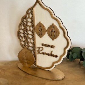 Pu&ograve; includere: Un calendario di conto alla rovescia in legno per il Ramadan con una finitura dorata. Il calendario presenta un design decorativo con una luna crescente e le parole "Giorni di Ramadan". I numeri 30 sono visualizzati sul calendario.