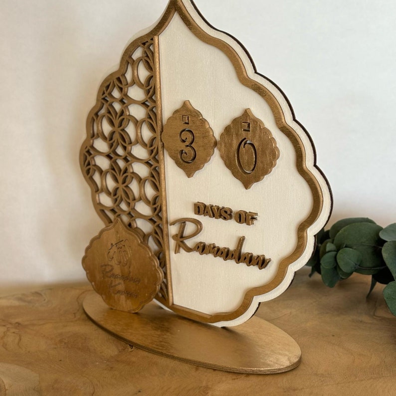 Pu&ograve; includere: Un calendario di conto alla rovescia in legno per il Ramadan con una finitura dorata. Il calendario presenta un design decorativo con una luna crescente e le parole "Giorni di Ramadan". I numeri 30 sono visualizzati sul calendario.