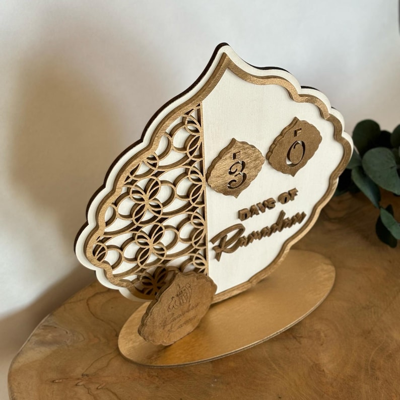 Pu&ograve; includere: Un calendario di conto alla rovescia per il Ramadan in legno con sfondo bianco e un design intricato tagliato al laser. Il calendario presenta un design ribaltabile con i numeri 30 e 0, e il testo "Giorni di Ramadan".
