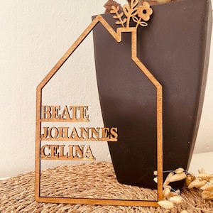 May include: Wooden house-shaped sign with the names "Beate", "Johannes", and "Celina" inscribed on it. The sign has a small flower design at the top.
